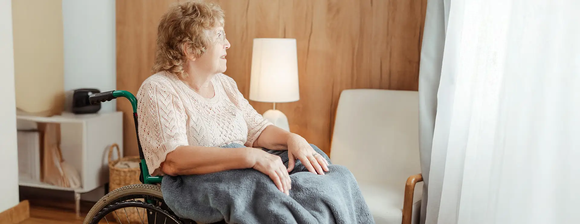 An older woman in a wheelchair, covered with a blue blanket, gazes out a window in the cozy, softly lit room at West Wind Village, with warm wooden walls and a gentle lamp nearby. An older woman in a wheelchair, covered with a blue blanket, gazes out a window in the cozy, softly lit room at West Wind Village, with warm wooden walls and a gentle lamp nearby.