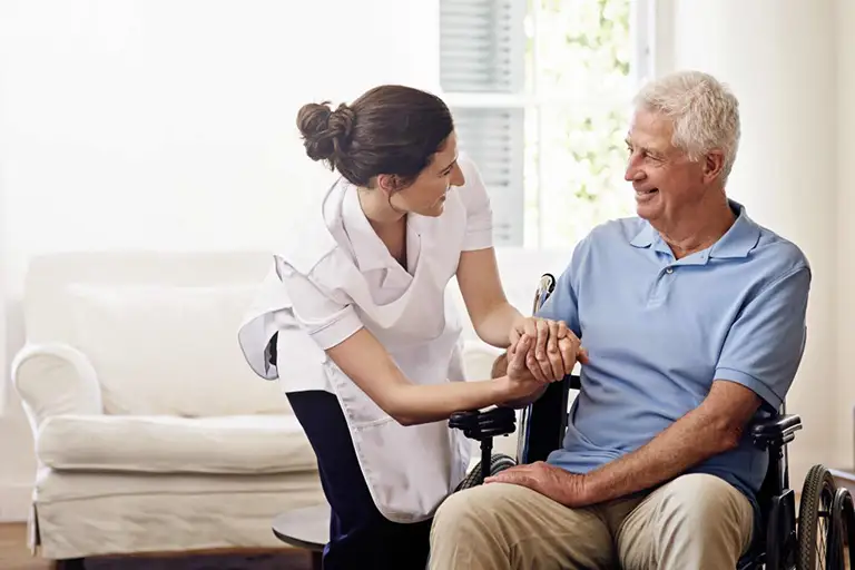 A nurse in a white uniform assists and smiles at an older man sitting in a wheelchair in a bright, home-like room. A nurse in a white uniform assists and smiles at an older man sitting in a wheelchair in a bright, home-like room.