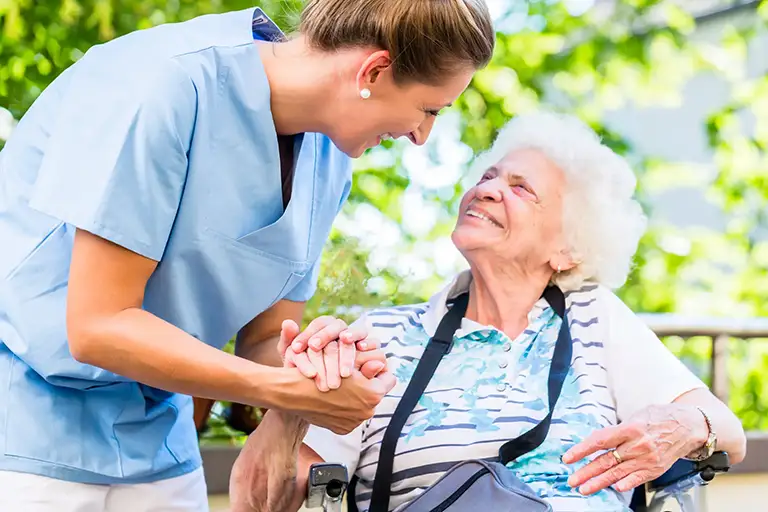 A nurse in blue scrubs from St. Francis Health Services holds hands with an elderly woman in a wheelchair outdoors, both smiling at each other.