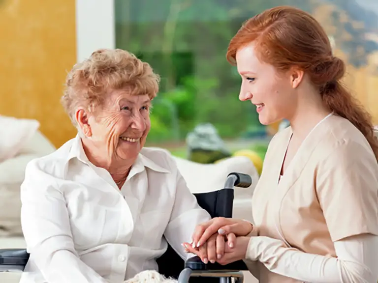 A smiling elderly woman in a wheelchair holds hands with a young caregiver, both looking at each other in a bright indoor setting at St. Francis Outpatient Therapy in Minnesota. A smiling elderly woman in a wheelchair holds hands with a young caregiver, both looking at each other in a bright indoor setting at St. Francis Outpatient Therapy in Minnesota.