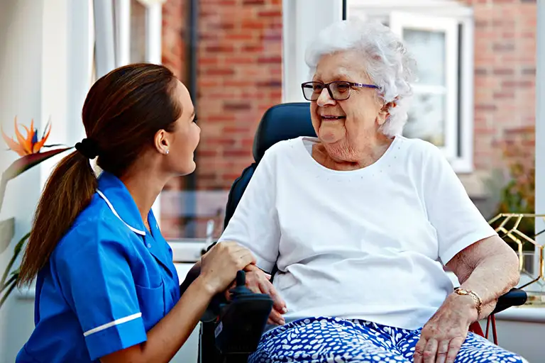 A nurse in blue scrubs sits beside and holds hands with an elderly woman in a wheelchair, both smiling inside a well-lit room at St. Francis Health Services. A nurse in blue scrubs sits beside and holds hands with an elderly woman in a wheelchair, both smiling inside a well-lit room at St. Francis Health Services.