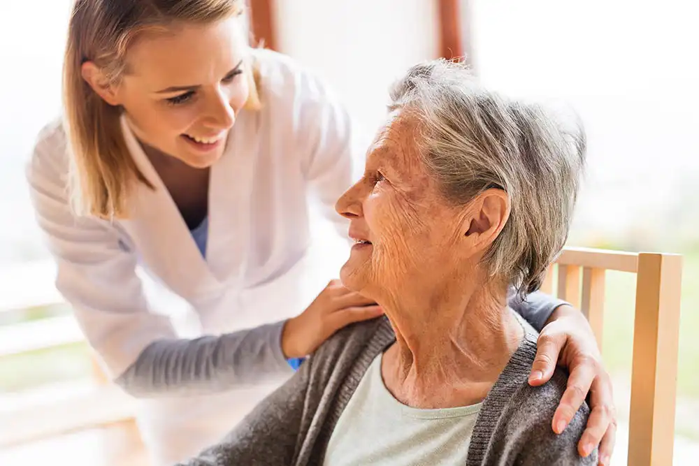 A young caregiver smiles and gently places her hand on the shoulder of an elderly woman sitting in a chair. A young caregiver smiles and gently places her hand on the shoulder of an elderly woman sitting in a chair.