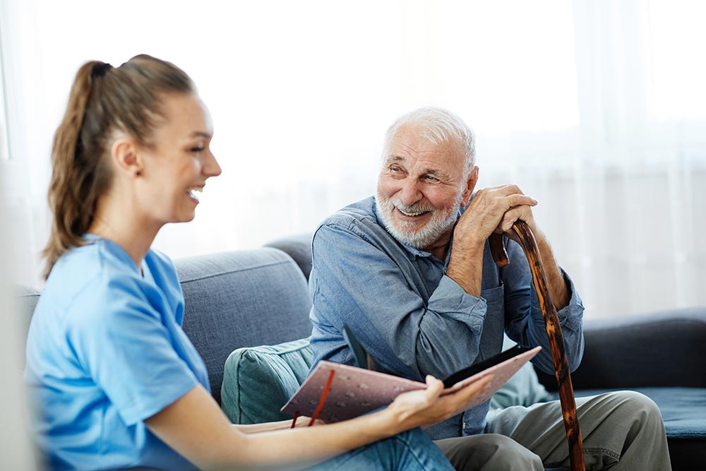 A caregiver in blue scrubs sits on a couch next to an elderly man with a cane, sharing a photo album and smiling together in a bright living room.