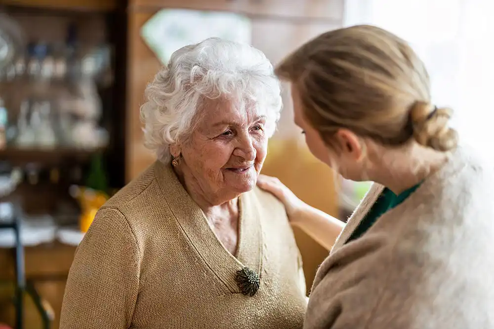 An elderly woman with white hair and a beige sweater smiles while a younger woman with light brown hair places a hand on her shoulder indoors. An elderly woman with white hair and a beige sweater smiles while a younger woman with light brown hair places a hand on her shoulder indoors.
