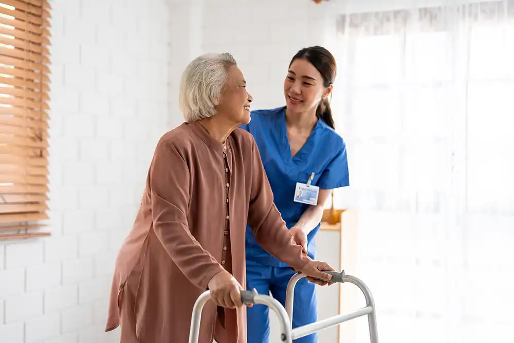An elderly woman using a walker is assisted by a healthcare worker in blue scrubs inside a bright room. An elderly woman using a walker is assisted by a healthcare worker in blue scrubs inside a bright room.