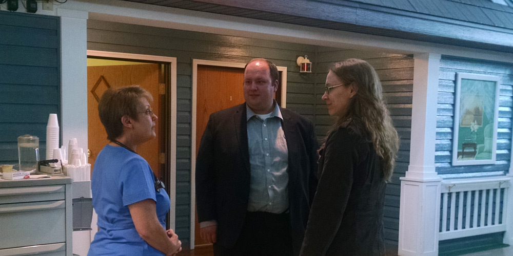 Three people stand indoors having a conversation; one woman wears scrubs, while the other two are in business attire, near a wall with doors and a framed picture. Three people stand indoors having a conversation; one woman wears scrubs, while the other two are in business attire, near a wall with doors and a framed picture.