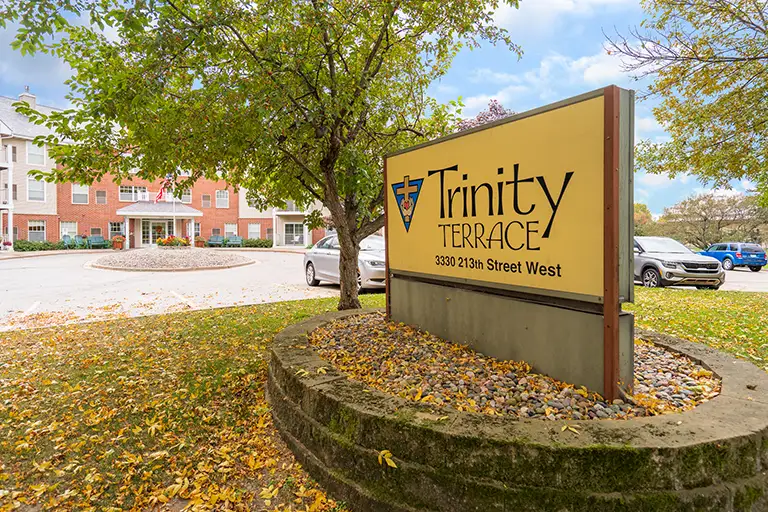 A large sign reading "Trinity Terrace, 3301 213th Street West" stands in front of a brick building, surrounded by autumn trees and parked cars.