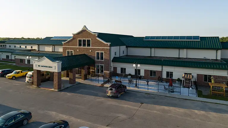 A large, two-story brick and white building with green roofs and solar panels, featuring a covered entrance and a parking lot with several cars. A large, two-story brick and white building with green roofs and solar panels, featuring a covered entrance and a parking lot with several cars.