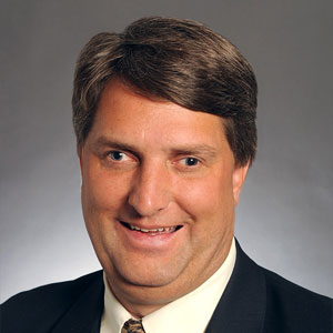 A man with short brown hair wearing a dark suit, light shirt, and patterned tie, smiling at the camera against a plain gray background. A man with short brown hair wearing a dark suit, light shirt, and patterned tie, smiling at the camera against a plain gray background.