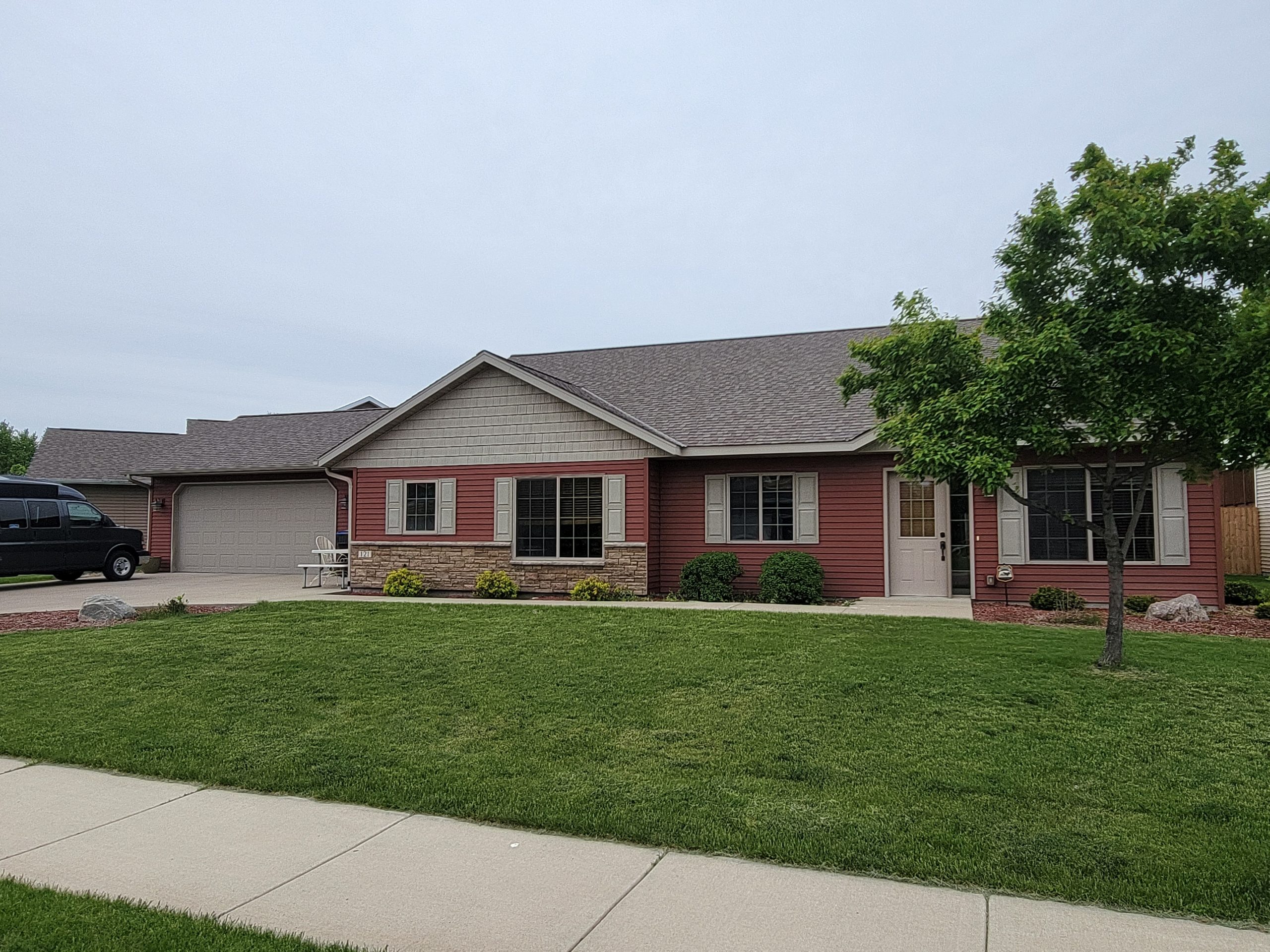 Single-story suburban house with red siding, stone accents, attached garage, large lawn, and a tree in the front yard on a cloudy day. Single-story suburban house with red siding, stone accents, attached garage, large lawn, and a tree in the front yard on a cloudy day.