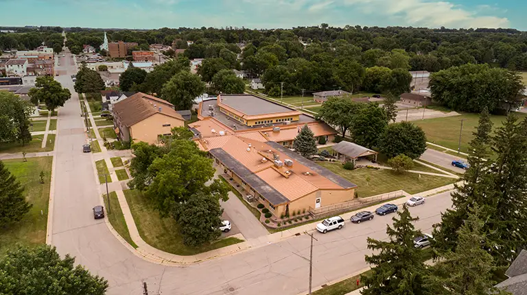 Aerial view of a school building surrounded by trees and residential streets with several parked cars lining the roads.