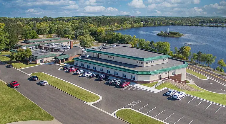 Aerial view of a modern building complex with green roofs, adjacent parking lots, and a lake surrounded by trees under a partly cloudy sky. Aerial view of a modern building complex with green roofs, adjacent parking lots, and a lake surrounded by trees under a partly cloudy sky.