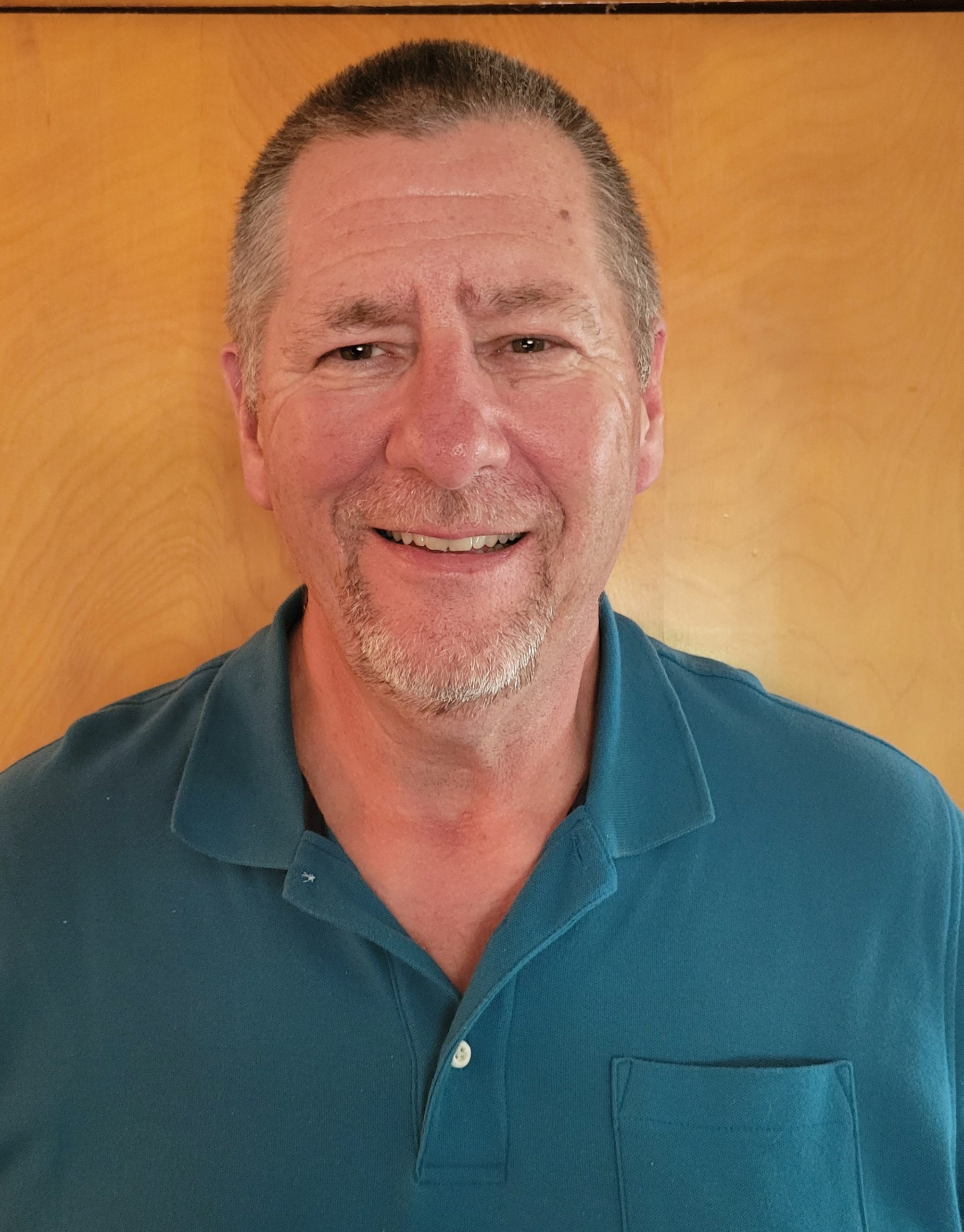 Curtis Bach, CFO, is a middle-aged man with short hair and a goatee, smiling in a teal polo shirt as he stands in front of a light wood background.