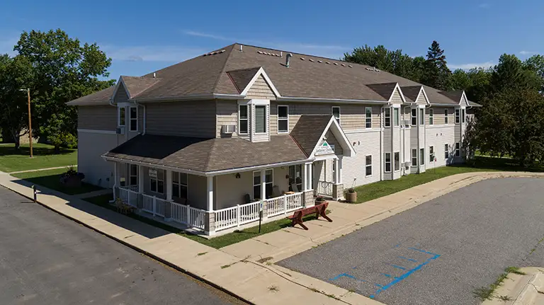Two-story residential building with a covered porch, surrounded by trees and a parking area with marked spaces, including a designated accessible spot.