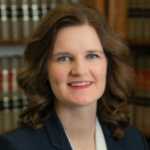 A woman with wavy brown hair, wearing a dark blazer and white top, smiles in front of bookshelves filled with legal books at St. Francis Health Services in Minnesota. A woman with wavy brown hair, wearing a dark blazer and white top, smiles in front of bookshelves filled with legal books at St. Francis Health Services in Minnesota.
