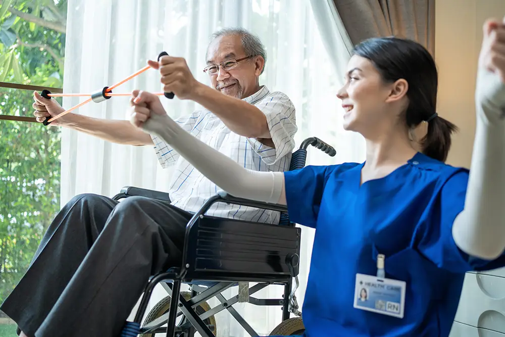 Elderly man in a wheelchair exercises with resistance bands, assisted by a female healthcare worker in blue scrubs. Elderly man in a wheelchair exercises with resistance bands, assisted by a female healthcare worker in blue scrubs.