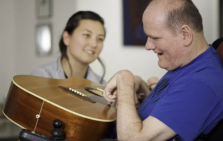 A man in a wheelchair strums a guitar while a woman nearby smiles at him, capturing the supportive atmosphere found at St. Francis Outpatient Therapy in Minnesota. A man in a wheelchair strums a guitar while a woman nearby smiles at him, capturing the supportive atmosphere found at St. Francis Outpatient Therapy in Minnesota.
