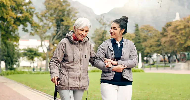 An elderly woman with a cane walks outdoors in a park, arm in arm with a younger woman. Both wearing jackets and deep in conversation, they embody the spirit of care found at St. Francis Outpatient Therapy, offered across Minnesota. An elderly woman with a cane walks outdoors in a park, arm in arm with a younger woman. Both wearing jackets and deep in conversation, they embody the spirit of care found at St. Francis Outpatient Therapy, offered across Minnesota.
