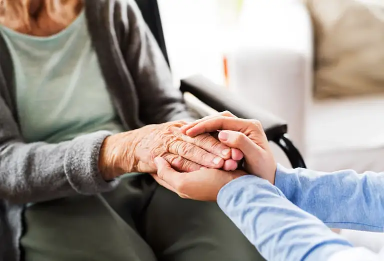 Close-up of a younger person gently holding the hands of an elderly person sitting in a wheelchair. Close-up of a younger person gently holding the hands of an elderly person sitting in a wheelchair.