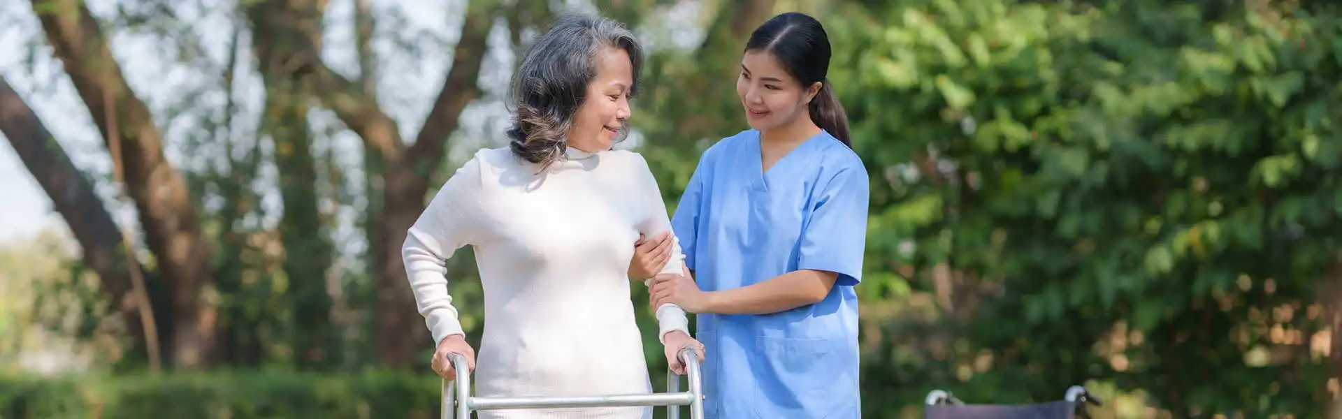 A senior woman using a walker is assisted by a caregiver in blue scrubs outdoors in a park-like setting. A senior woman using a walker is assisted by a caregiver in blue scrubs outdoors in a park-like setting.