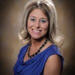 A woman with light brown, shoulder-length hair wearing a blue sleeveless top and a black beaded necklace, smiling at the camera against a brown gradient background, representing Outpatient Therapy in Minnesota at St. Francis Health Services. A woman with light brown, shoulder-length hair wearing a blue sleeveless top and a black beaded necklace, smiling at the camera against a brown gradient background, representing Outpatient Therapy in Minnesota at St. Francis Health Services.