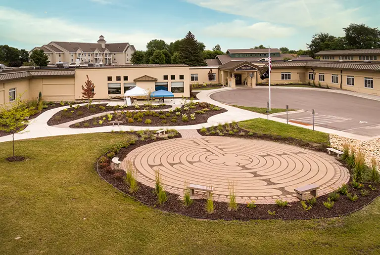 A landscaped courtyard with a circular walking labyrinth in the foreground leads to a large building with a peaked roof and multiple entrances. A landscaped courtyard with a circular walking labyrinth in the foreground leads to a large building with a peaked roof and multiple entrances.