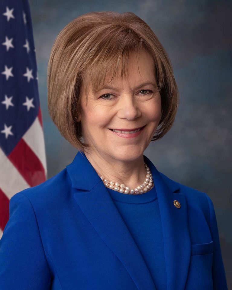 A woman in a blue blazer and pearl necklace poses in front of a US flag and a cloudy blue backdrop. A woman in a blue blazer and pearl necklace poses in front of a US flag and a cloudy blue backdrop.